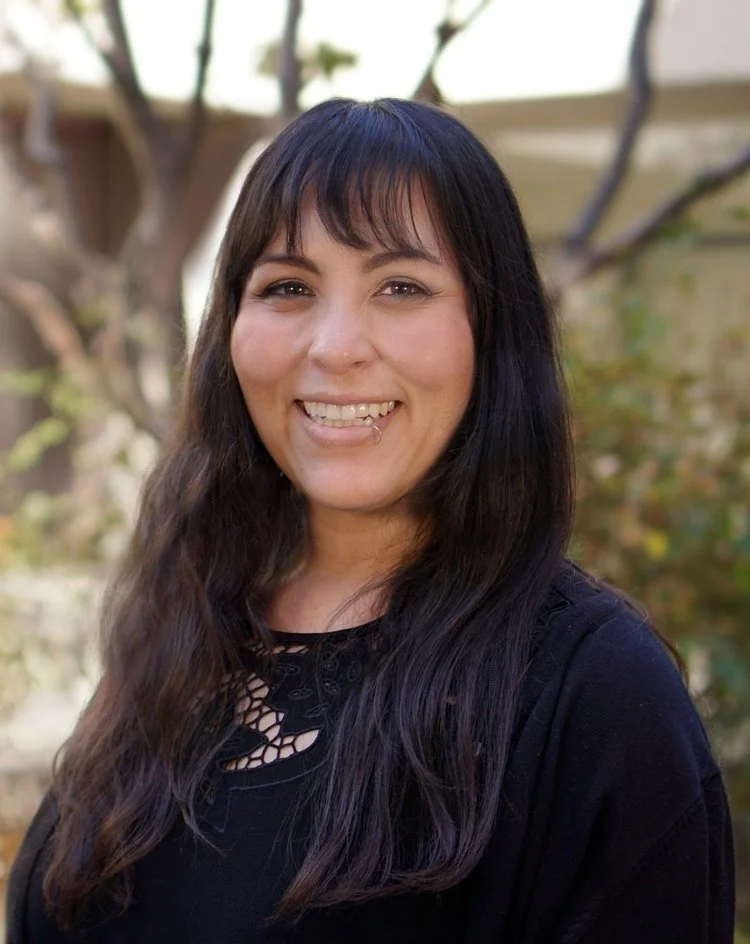 A woman with long dark hair, wearing a black top with intricate cut-out design, standing outdoors. She is smiling and has a friendly expression with a blurred garden background.