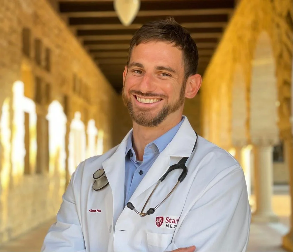 A male doctor with short dark hair and a beard wearing a white coat and blue shirt, smiling in a hallway with stone walls and windows.