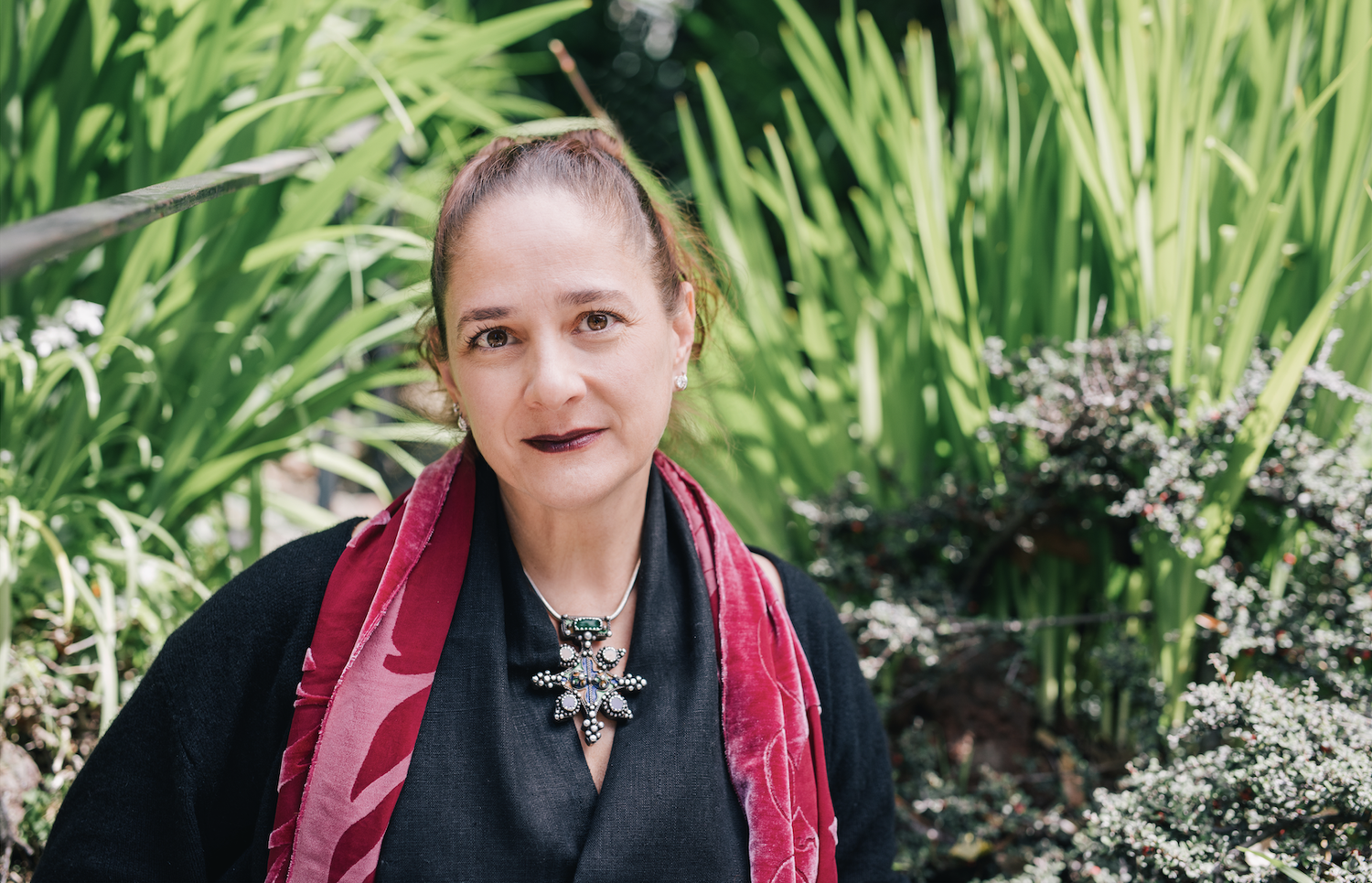 A woman with brown hair in a ponytail, wearing a dark top and a red scarf, standing outdoors with lush green plants in the background.