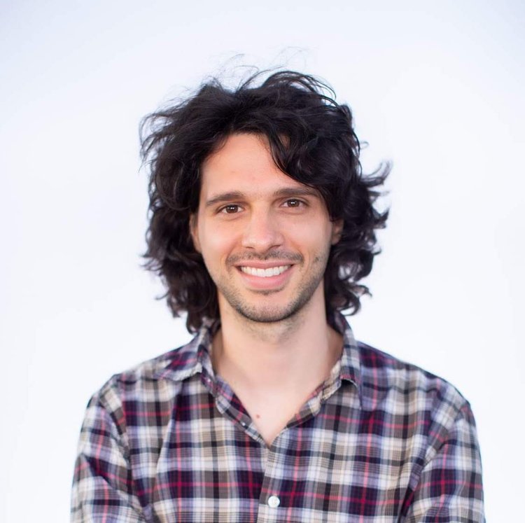 A young man with long, curly black hair, a light beard, and a bright smile wearing a plaid shirt, standing against a plain white background.
