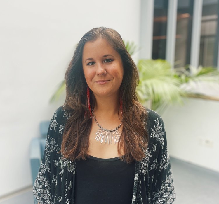 A woman with long brown hair, wearing a black top, black and white patterned cardigan, a silver statement necklace, and red earrings, standing indoors near a large window with plants in the background.