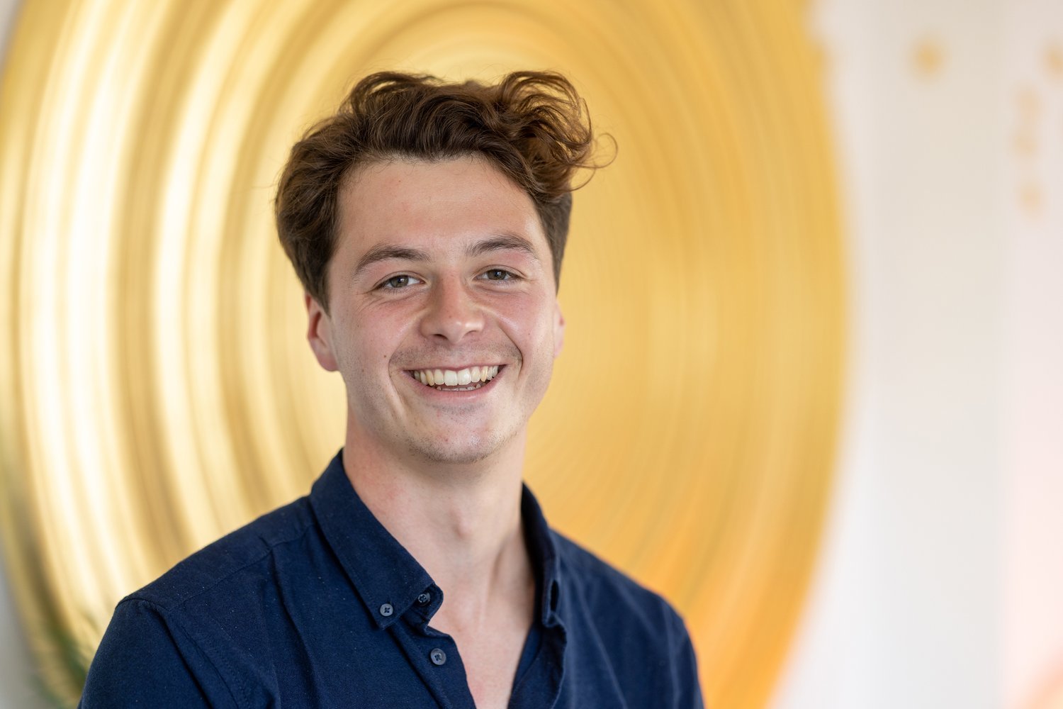 A smiling young man with brown hair wearing a navy blue button-up shirt, standing in front of a blurred yellow circular artwork or decoration.