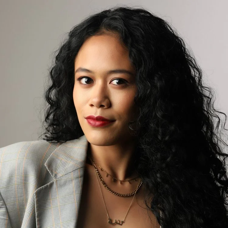 Portrait of a woman with curly black hair, wearing a plaid blazer and layered gold necklaces, looking confidently at the camera.