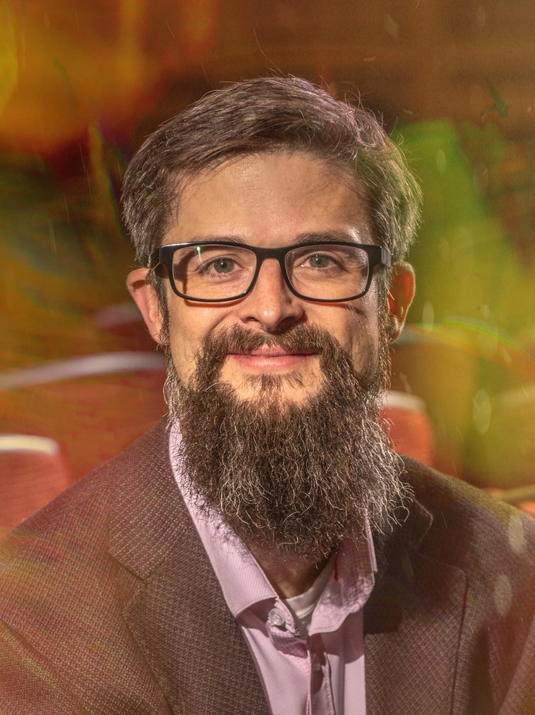 Portrait of a man with glasses and a full beard, wearing a suit jacket and a light pink shirt, smiling in a colorful, blurred background.