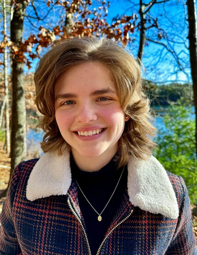 A young woman with short wavy hair smiling outdoors with trees and a body of water in the background.