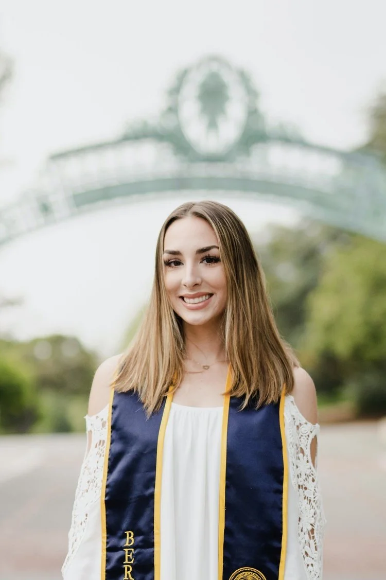 Young woman in a graduation gown and stole smiling outdoors, with an arch and trees in the background.