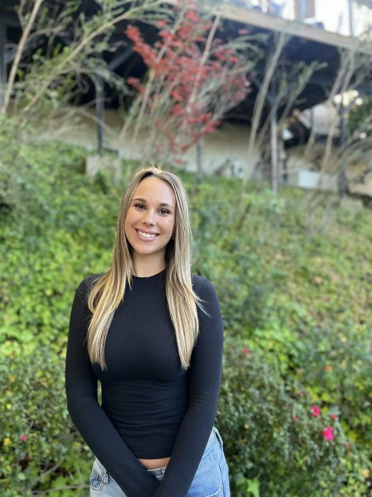 Young woman with long blonde hair, wearing a black long-sleeve shirt and jeans, standing outdoors in front of green shrubbery and a tree with red leaves.