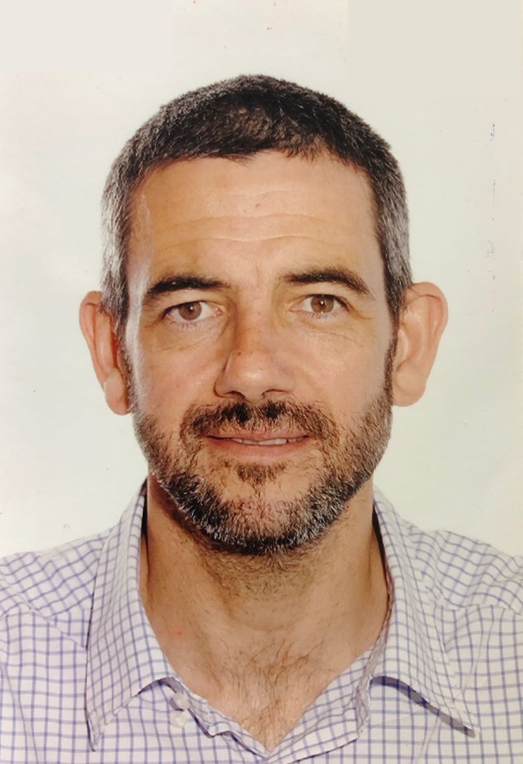 Headshot of a man with short, dark hair, stubble, wearing a light checkered shirt, smiling softly, against a plain white background.