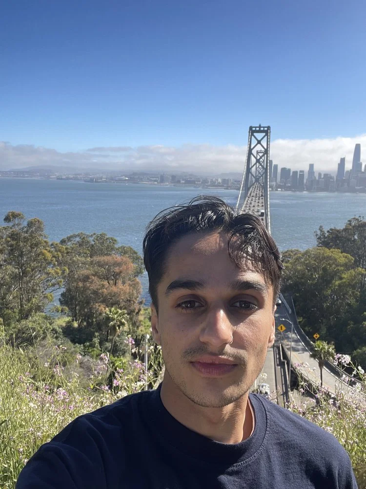 A young man taking a selfie outdoors with a suspension bridge, water, and city skyline in the background on a clear day.