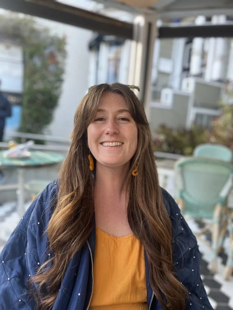 A smiling woman with long brown hair wearing a navy blue jacket and yellow earrings, sitting outside on a patio with a background of house windows, plants, and patio furniture.