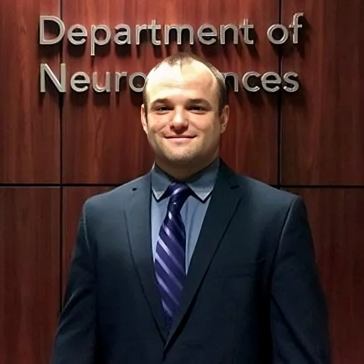 A young man in a suit and tie standing in front of a wooden wall with a sign that reads 'Department of Neurosciences'.