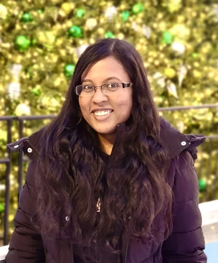 A woman with long, curly hair and glasses smiling at the camera, standing in front of a decorated Christmas tree with green and gold ornaments.