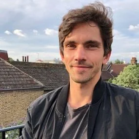 A young man with brown hair and a slight smile, wearing a black jacket and a gray shirt, standing outdoors with rooftops and trees in the background