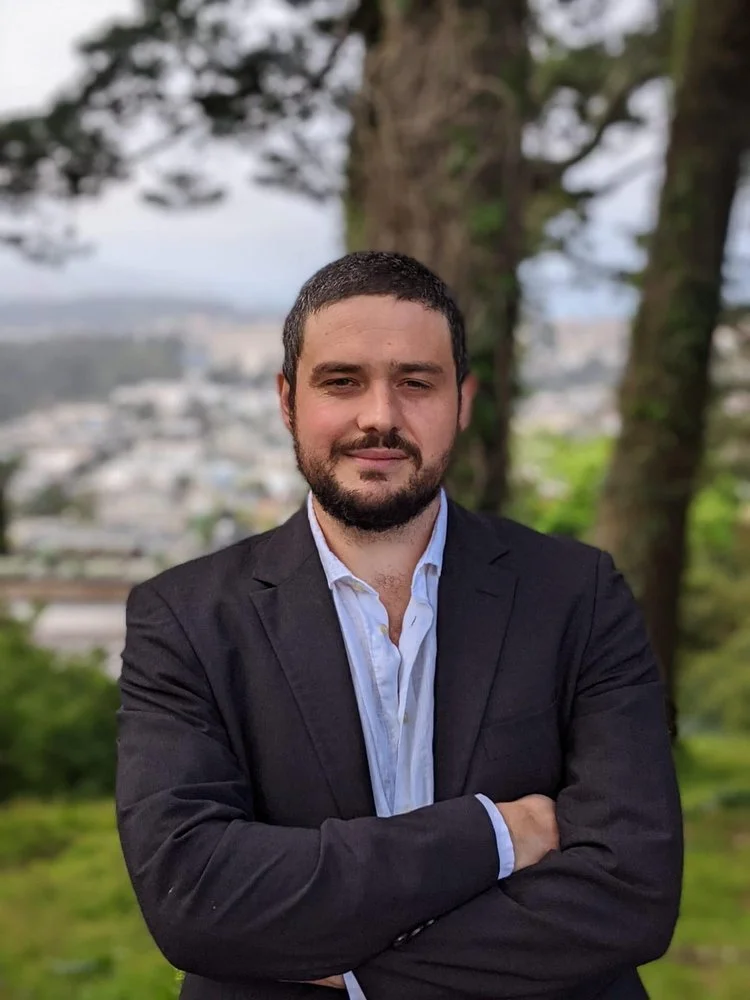 Man in a black blazer and light blue shirt standing outdoors with cross-armed pose, trees and cityscape in the background.