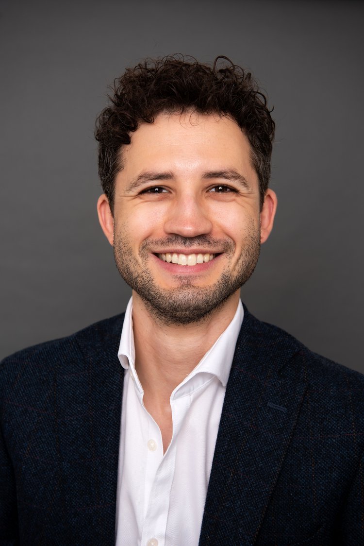 Portrait of a smiling man with curly dark hair, wearing a white shirt and a dark blazer, against a gray background.