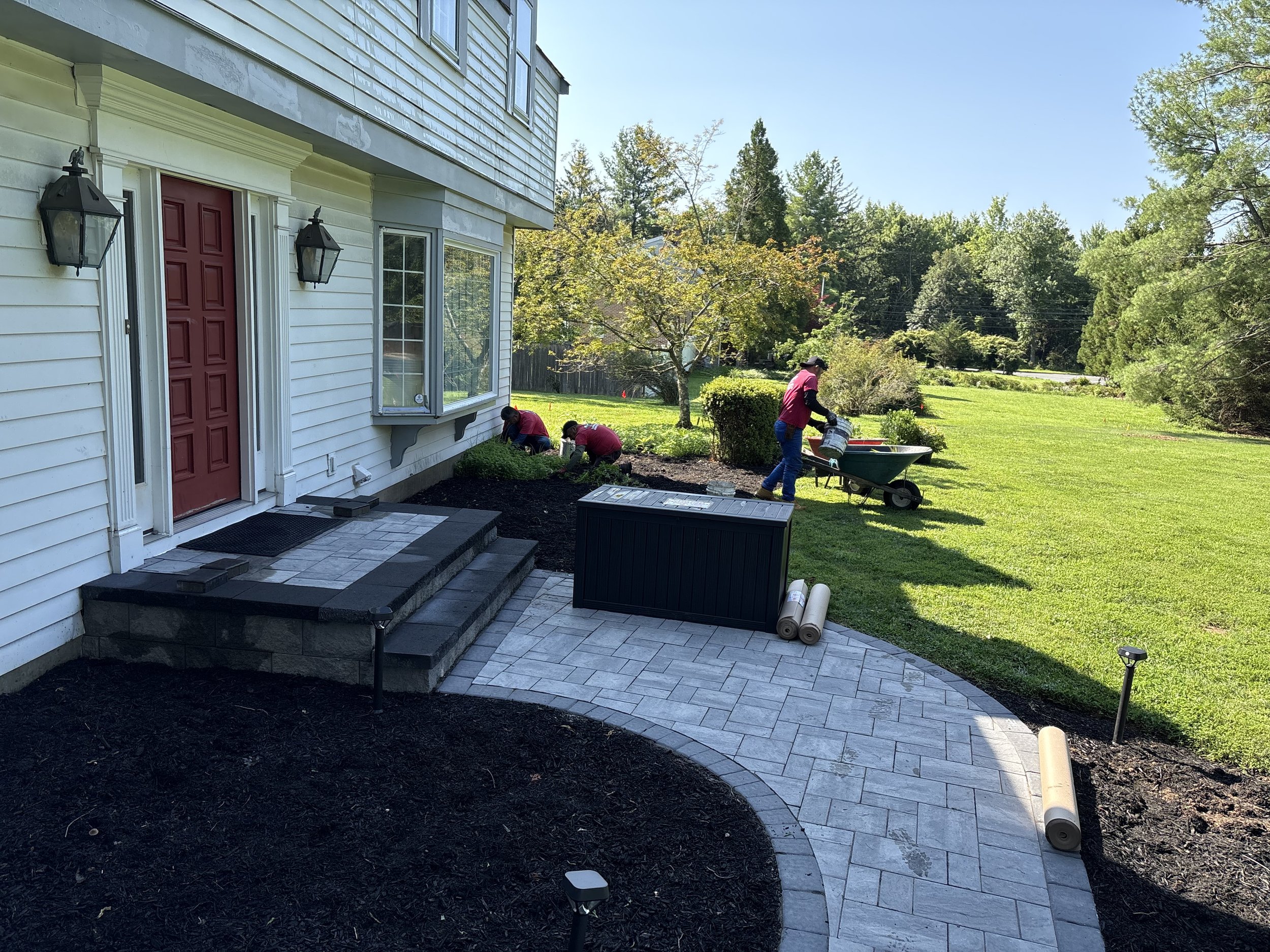 People planting shrubs and flowers in a landscaped garden bed with a rock border, trees, and a backyard fence on a sunny day.