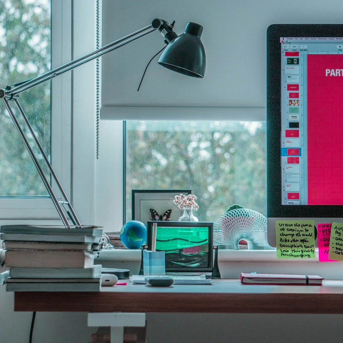 A cluttered desk with a large monitor showing a red presentation slide, a stack of papers, a small globe, a glass of water, a decorative shell, sticky notes, a monitor arm lamp, and a window with a view of trees outside.