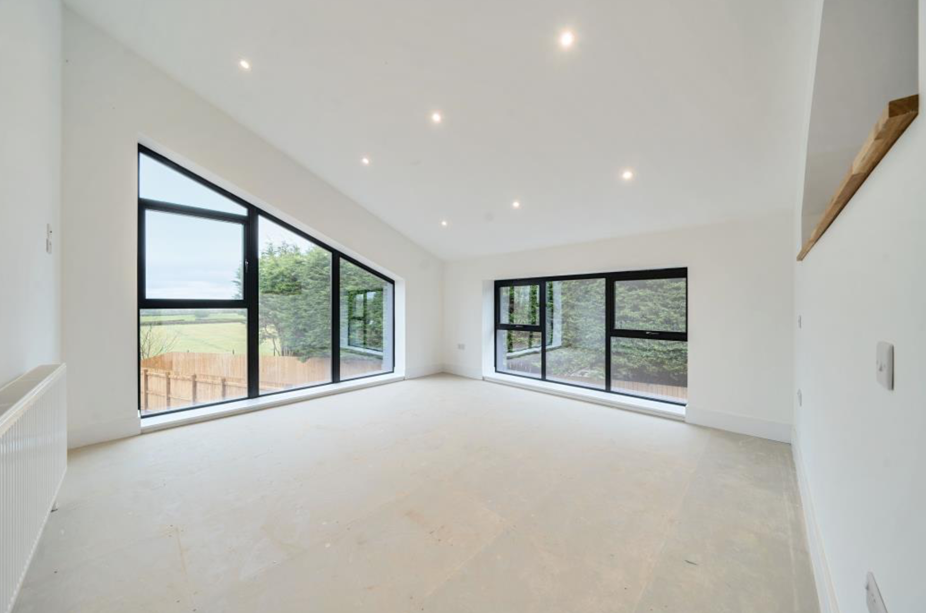 Empty modern room with large triangular and rectangular windows, white walls, angled ceiling, recessed lighting, and view of outdoor trees and grassy field.