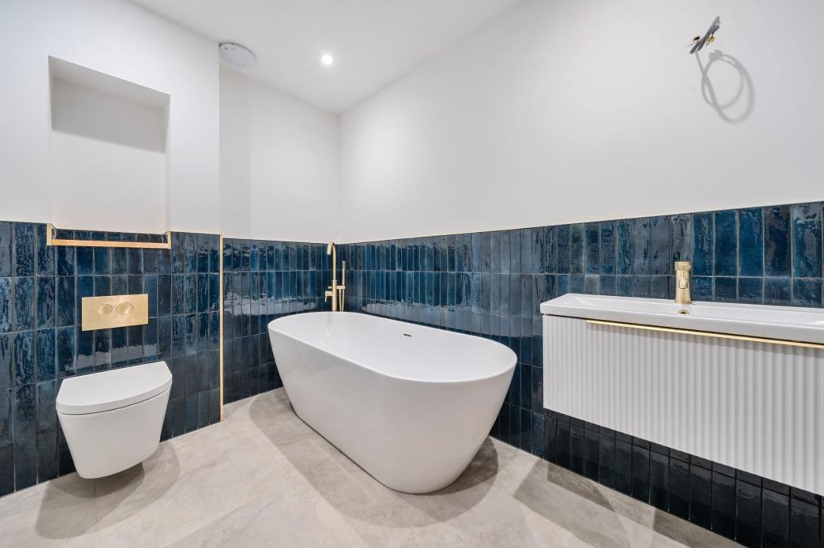 Modern bathroom with freestanding bathtub, wall-mounted toilet, and vanity sink; blue tiled walls and beige floor tiles.