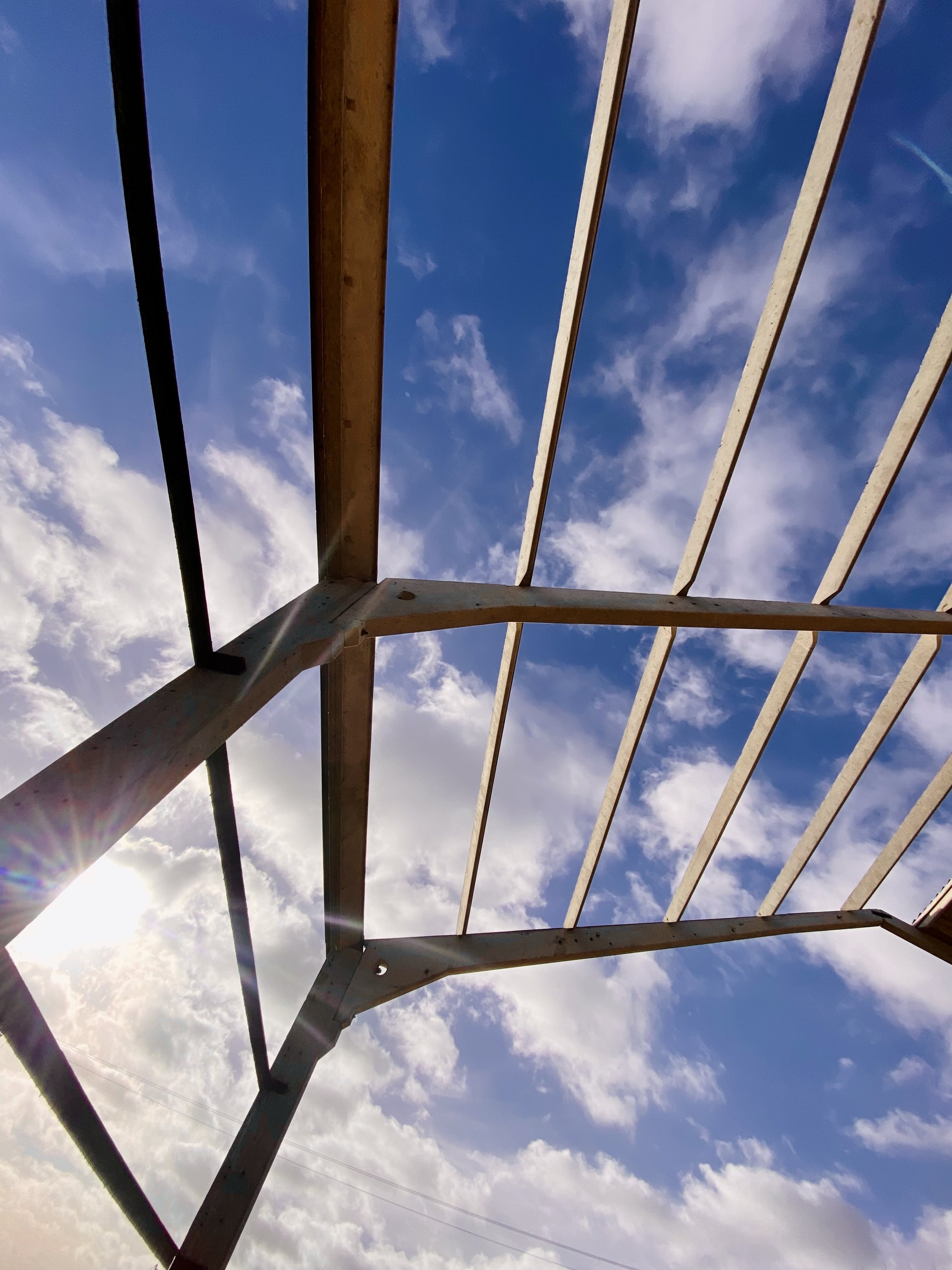 Metal framework of a roof or pergola against a blue sky with clouds and sunlight streaming through.