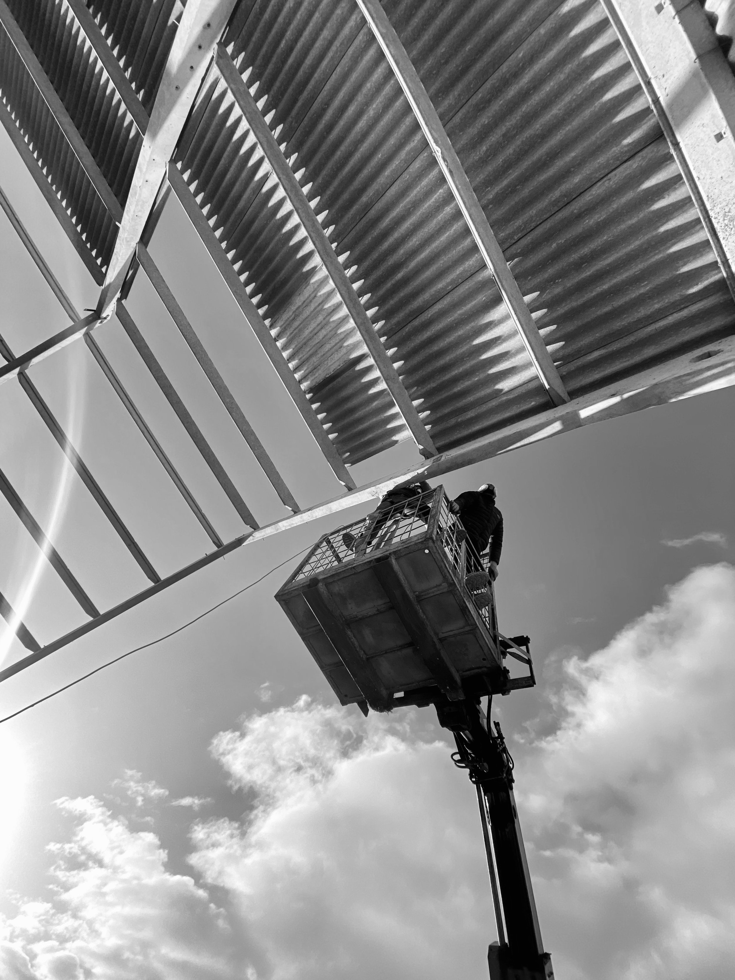 Worker on a lift under corrugated metal structure, viewed from below, against a cloudy sky in black and white.