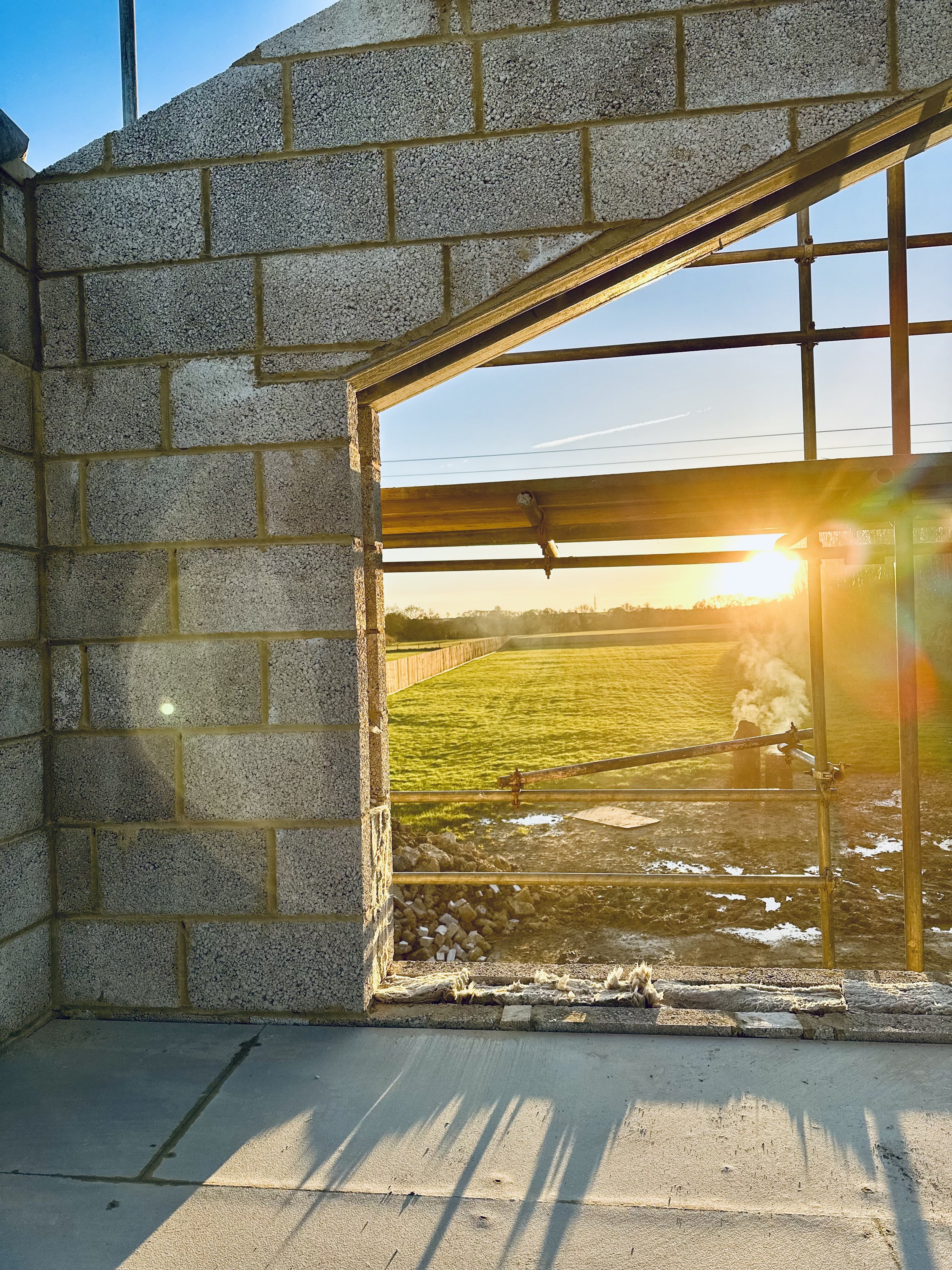 Sunset view through an unfinished brick wall window opening, overlooking a construction site and grassy field.