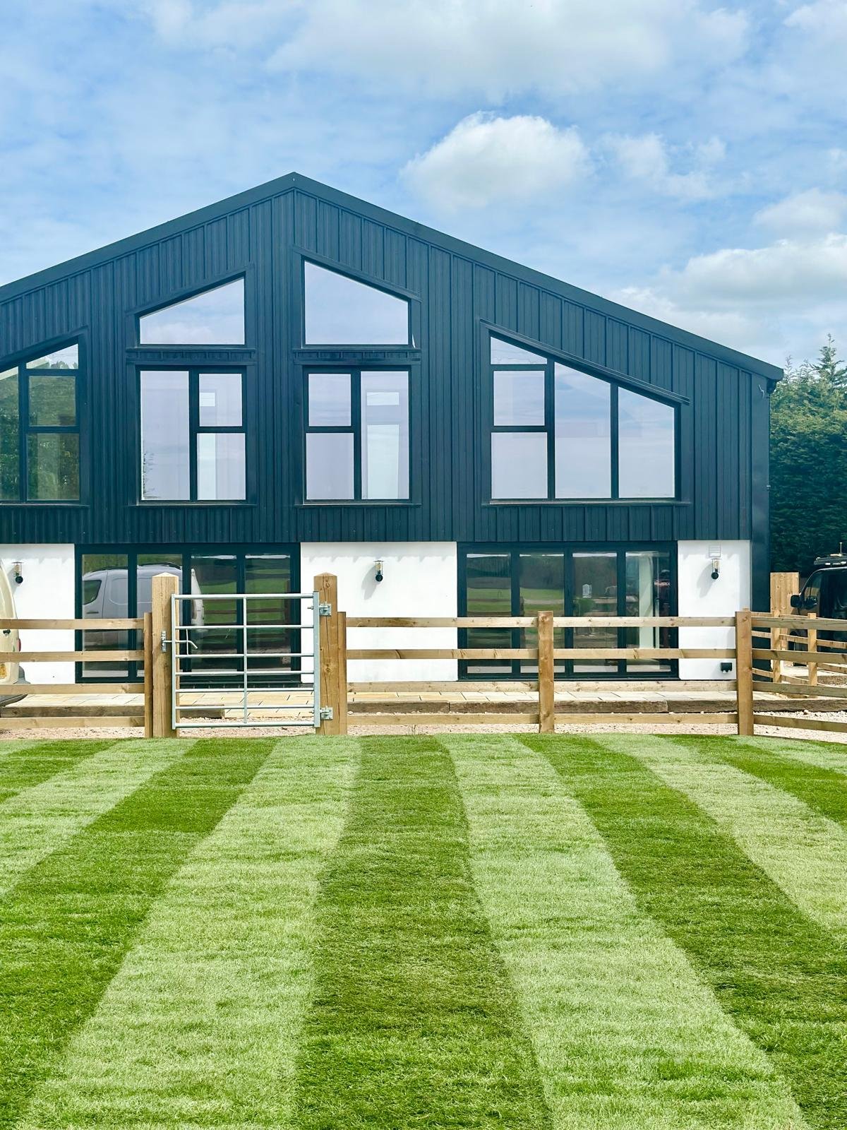 Modern black and white barn-style house with large windows, wooden fence, and freshly mowed lawn under a blue sky with clouds.