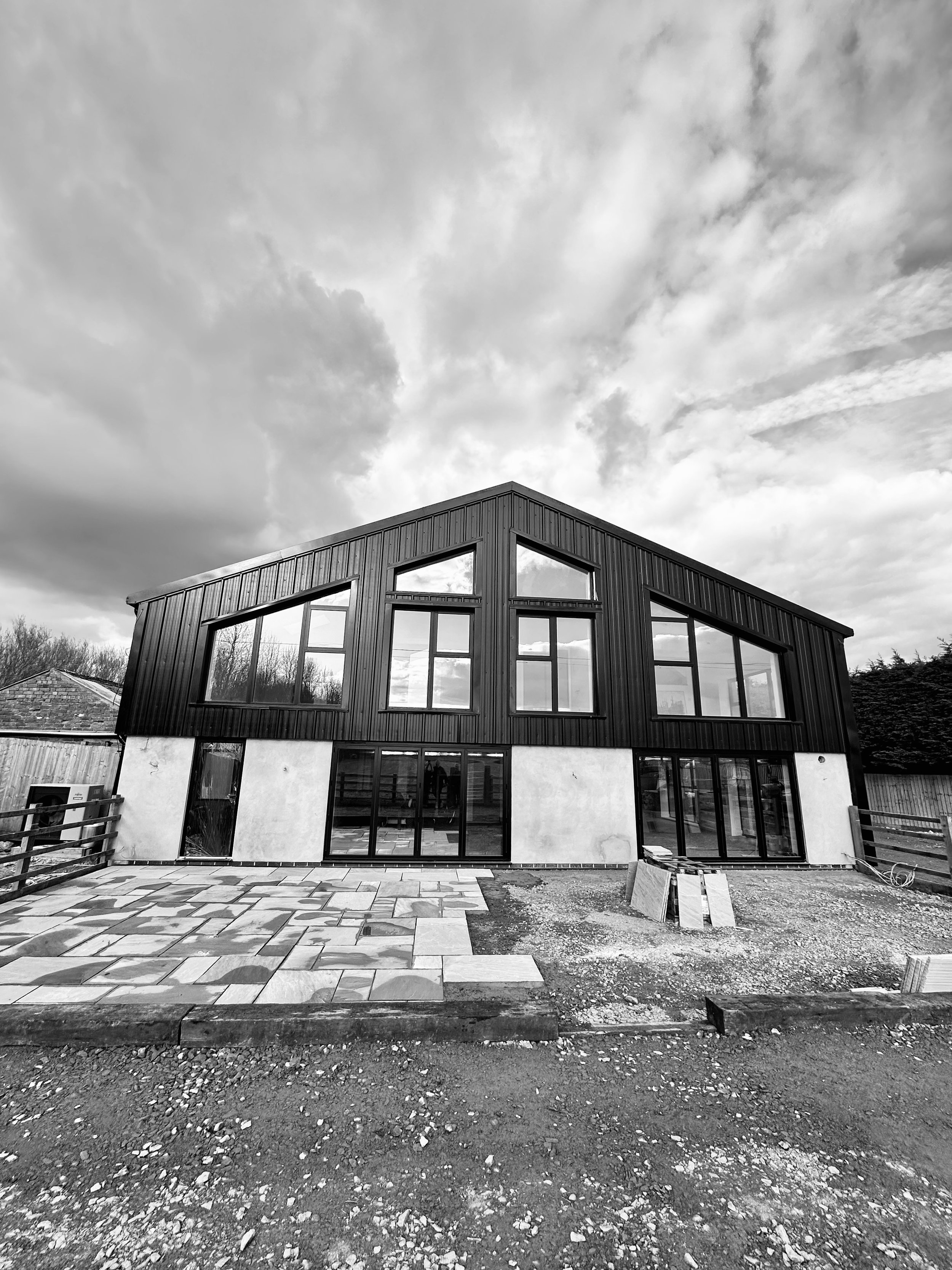 Black and white image of a modern building with large windows and a patio area in development, under a cloudy sky.