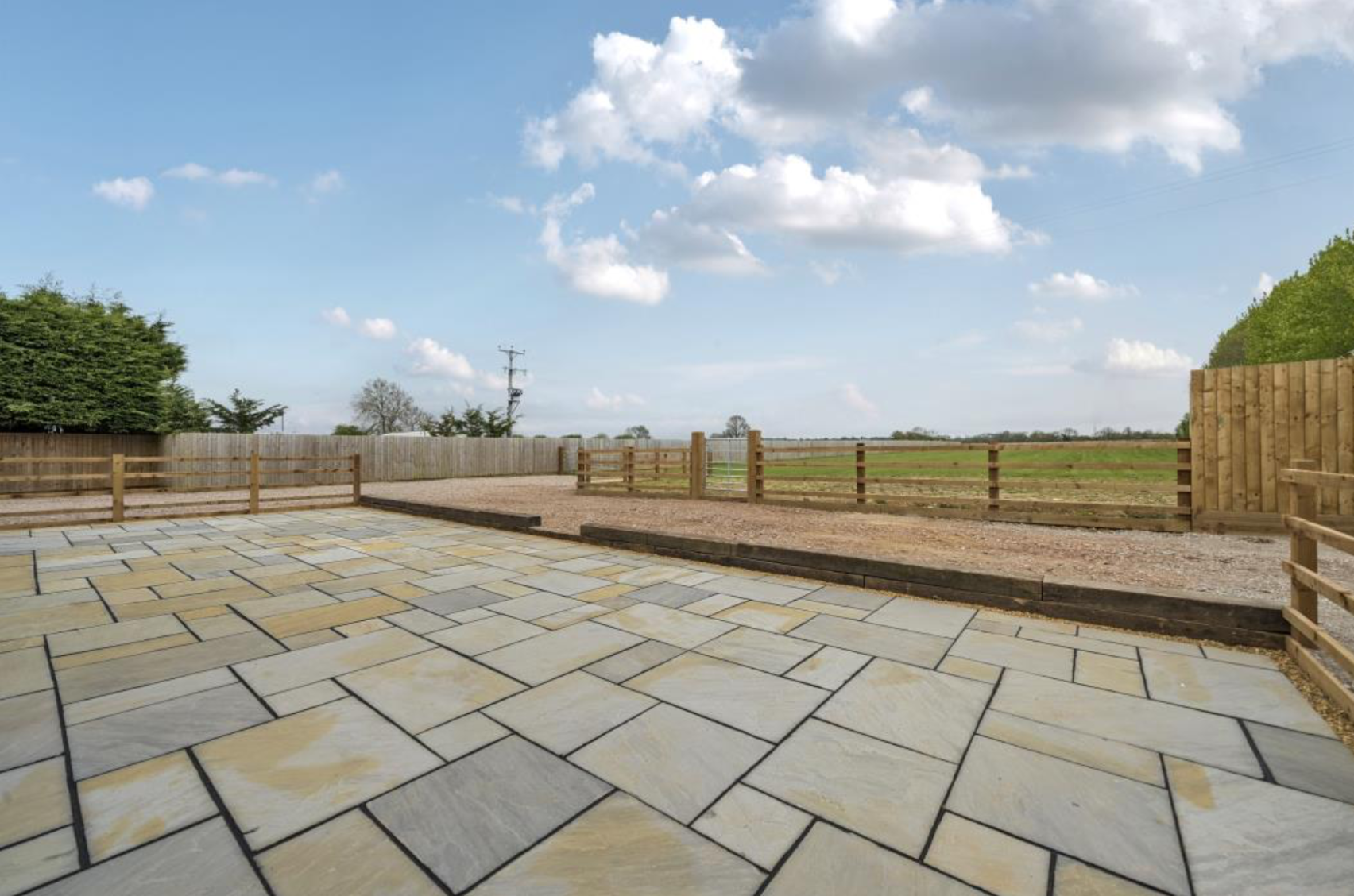 Paved patio area overlooking a grassy field with wooden fencing and trees under a partly cloudy sky.