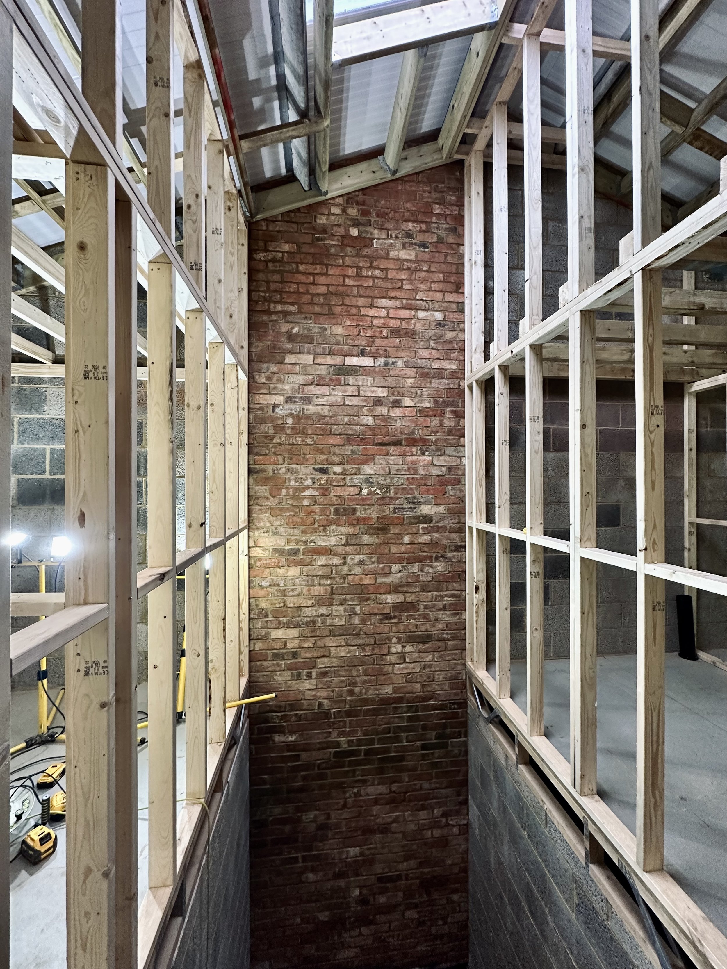 Interior view of a construction site with exposed wooden framing, a brick wall, and overhead roofing. Construction equipment and tools are visible on the floor.