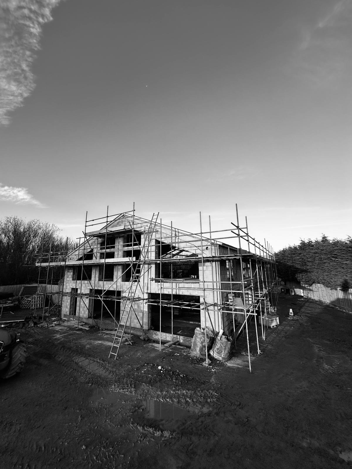 Black and white image of a partially constructed building with scaffolding, a ladder, and construction materials around it. The sky is mostly clear with some clouds, and trees are visible in the background.