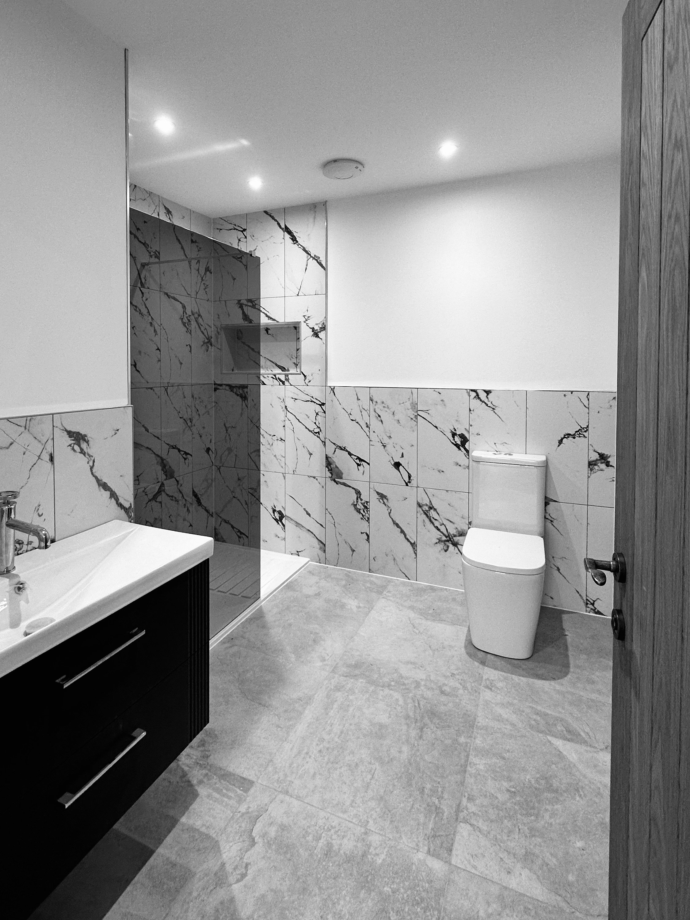 Modern bathroom with marble-tiled walls and a walk-in shower, featuring a sink and cabinet on the left and a toilet on the right.