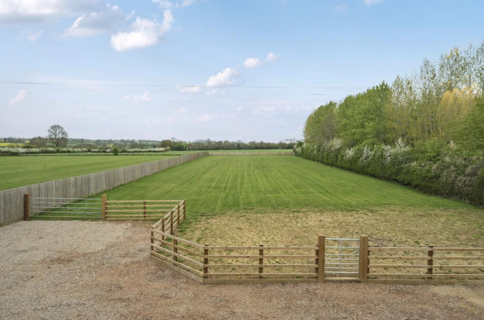 Aerial view of a large grass field bordered by a wooden fence and trees under a blue sky with clouds.