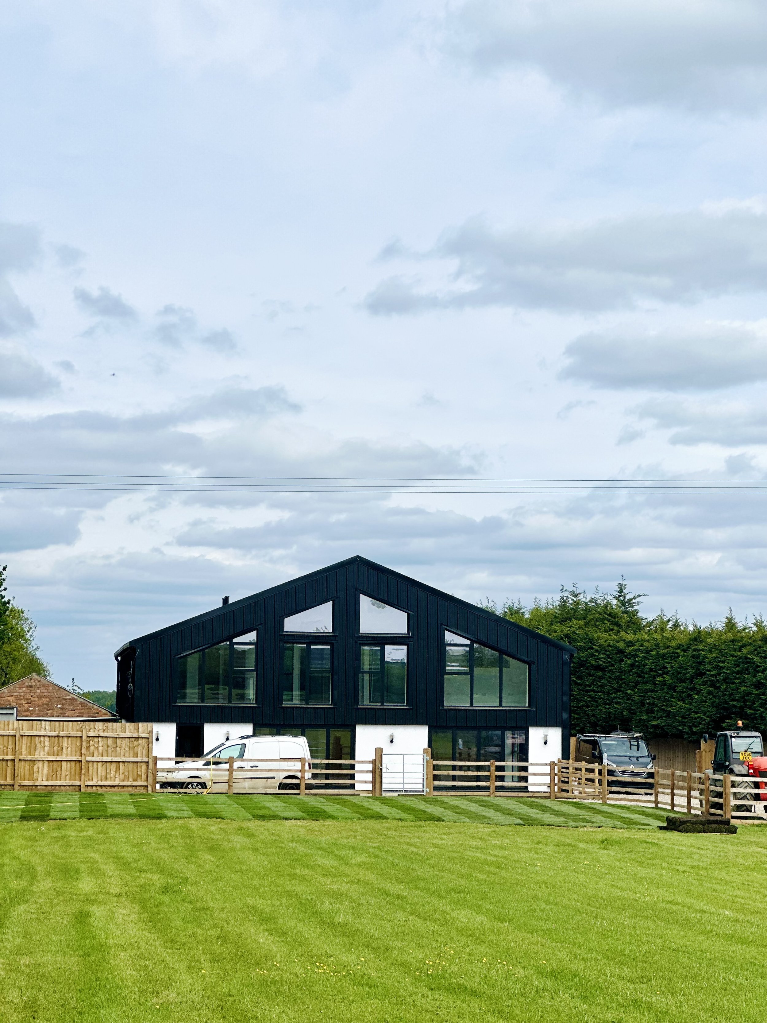 Modern barn-style house with large windows and dark exterior, surrounded by a fenced lawn, vehicles parked nearby, under a cloudy sky.