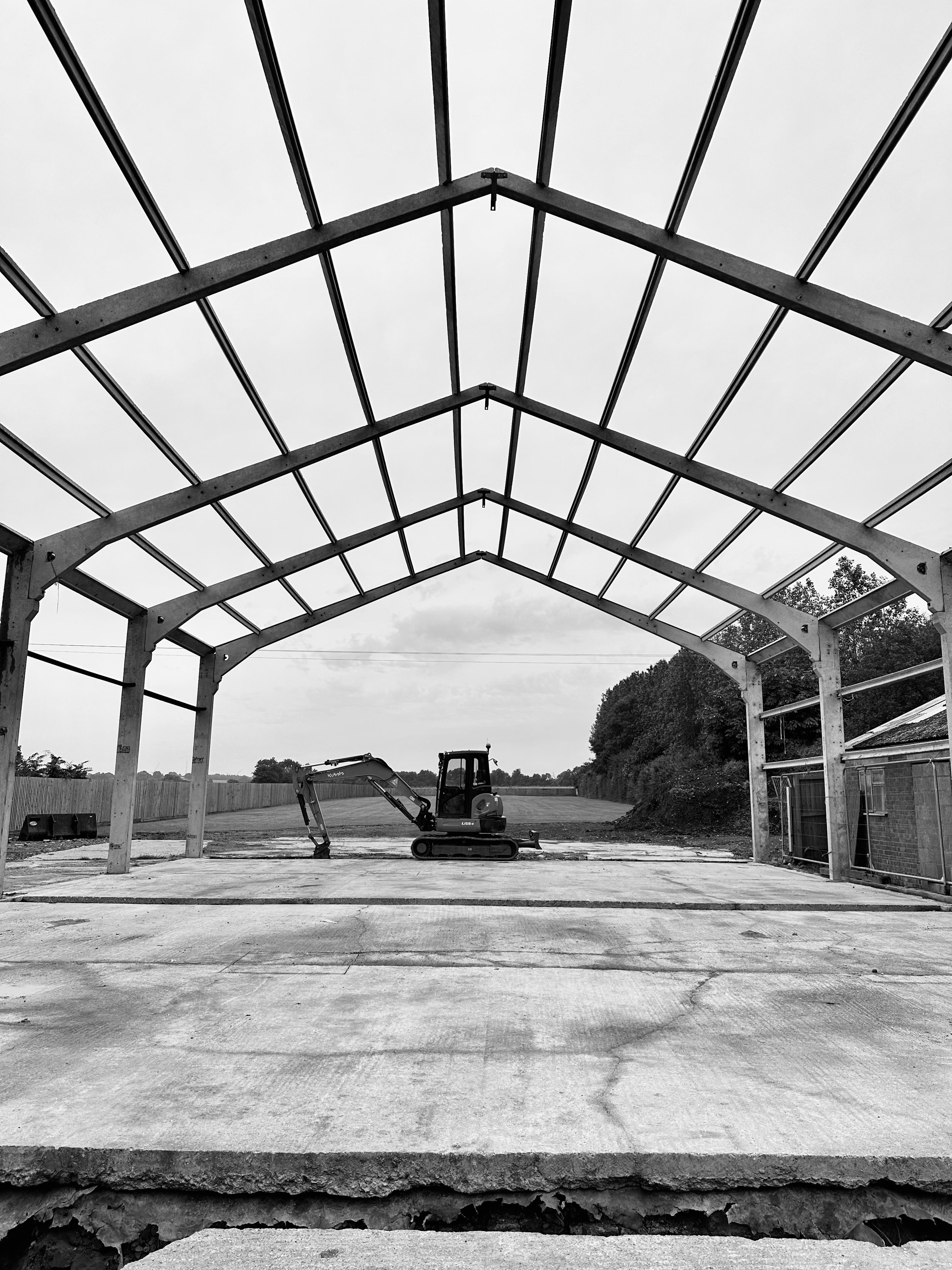 Black and white image of an incomplete building frame with a mini excavator on a concrete floor, surrounded by open land and trees.