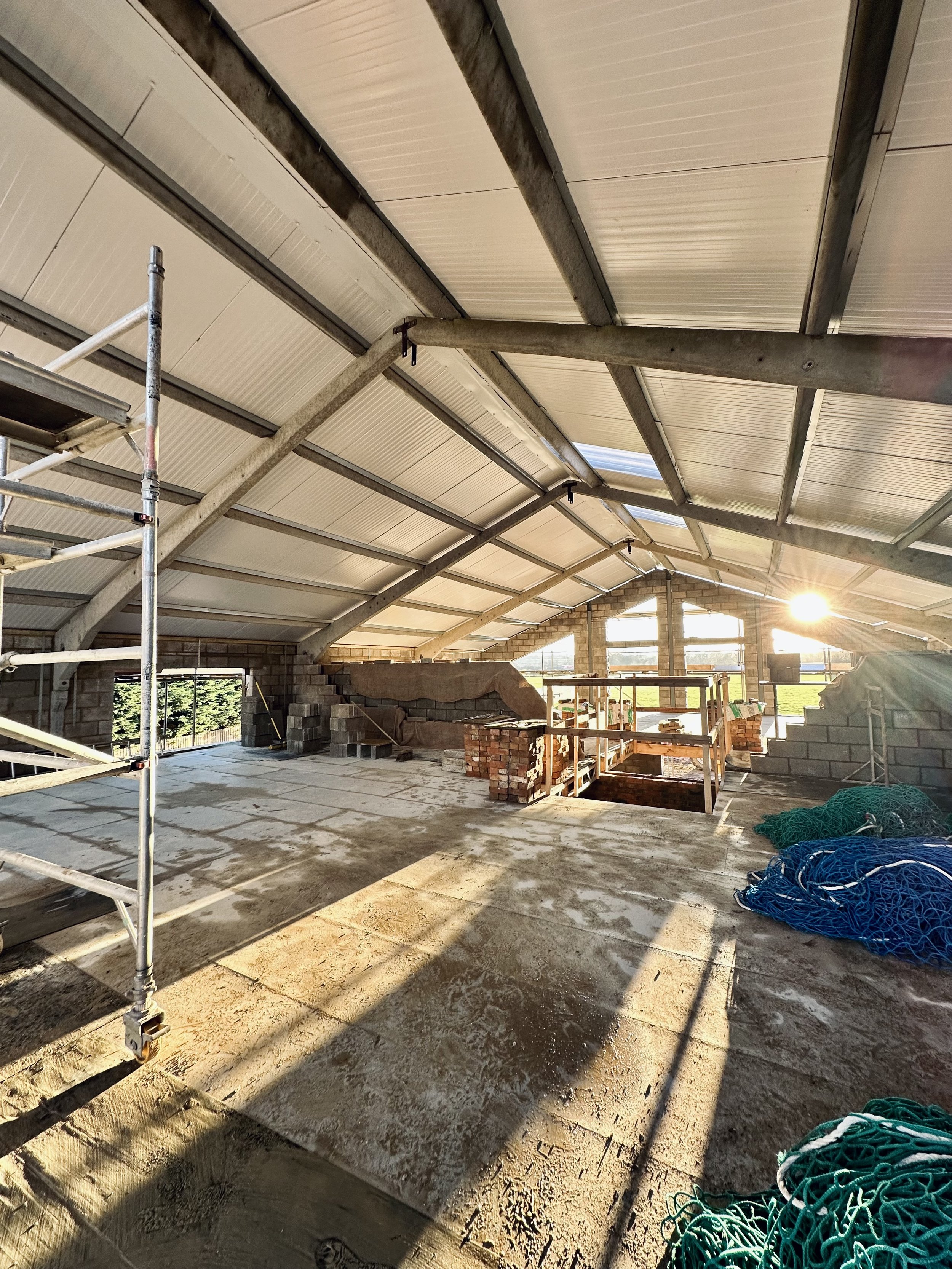 Interior view of a construction site with a partially completed roof, scaffolding, building materials, and sunlight streaming in.