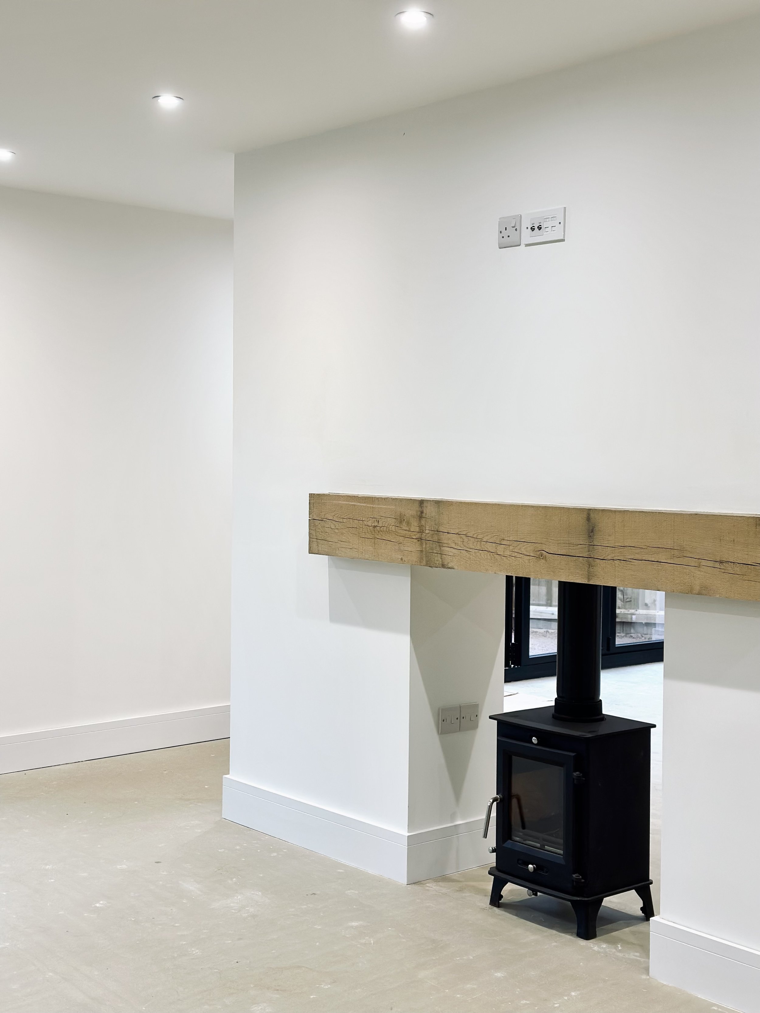 Minimalist interior with a small black wood stove set against a white wall, under a wooden mantelpiece. Recessed ceiling lights illuminate the room with bare floors.