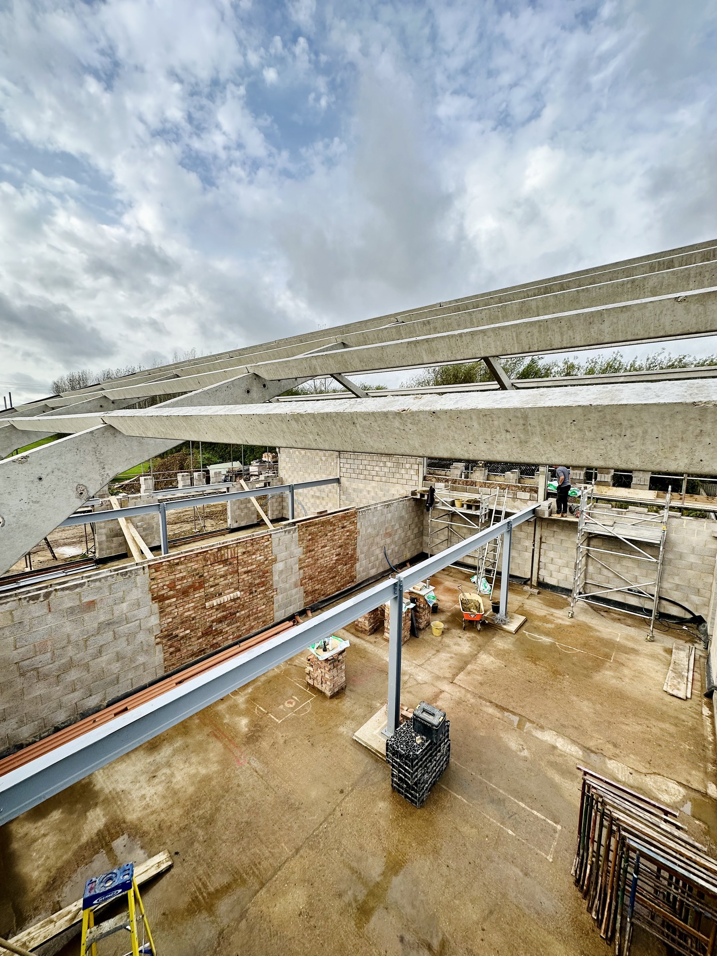 Construction site with partially built brick walls, steel beams, scaffolding, and construction materials on a concrete floor under a cloudy sky.