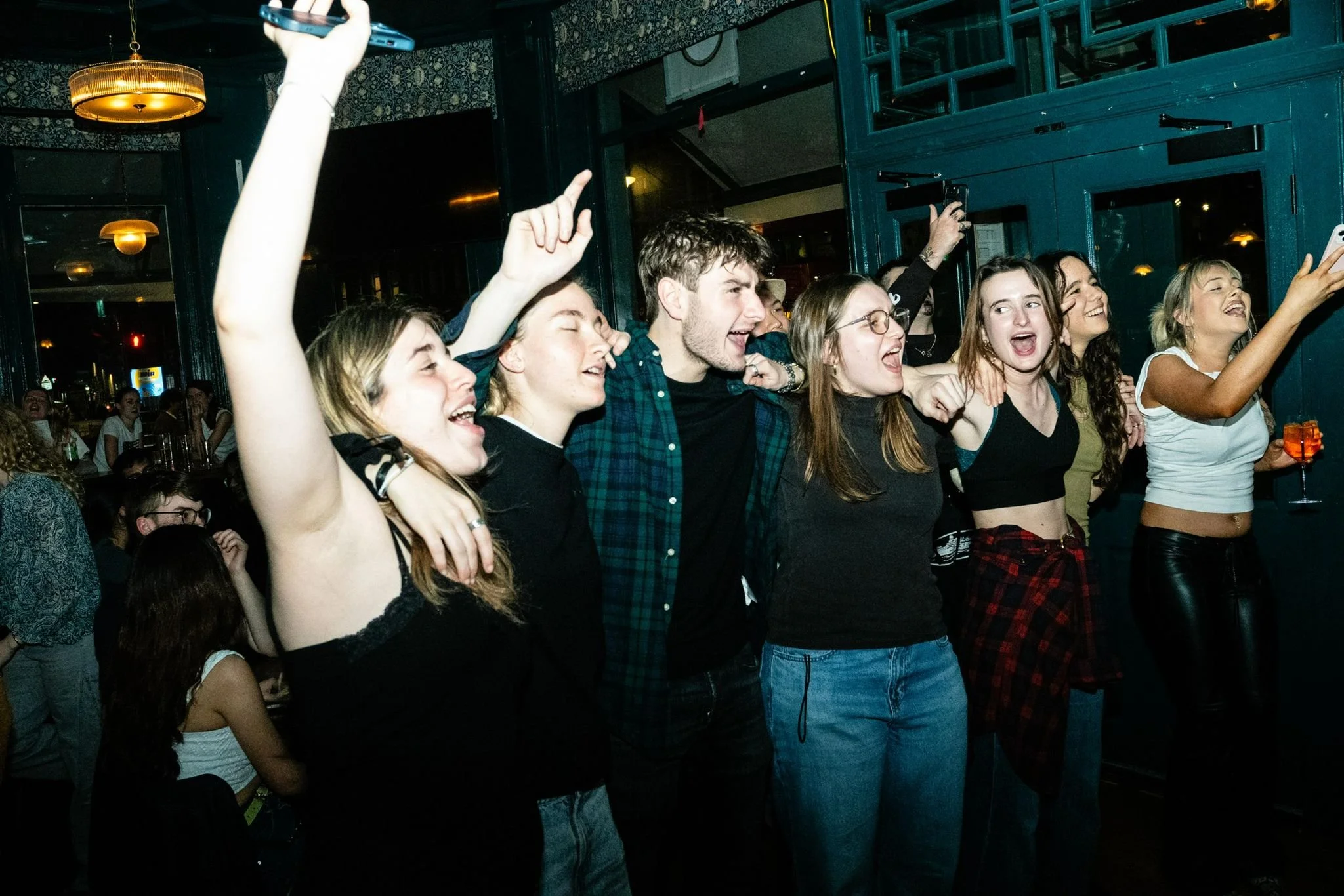 Group of young people cheerfully singing and dancing at a party or concert, some with arms around each other, in a dark indoor setting with warm lighting.