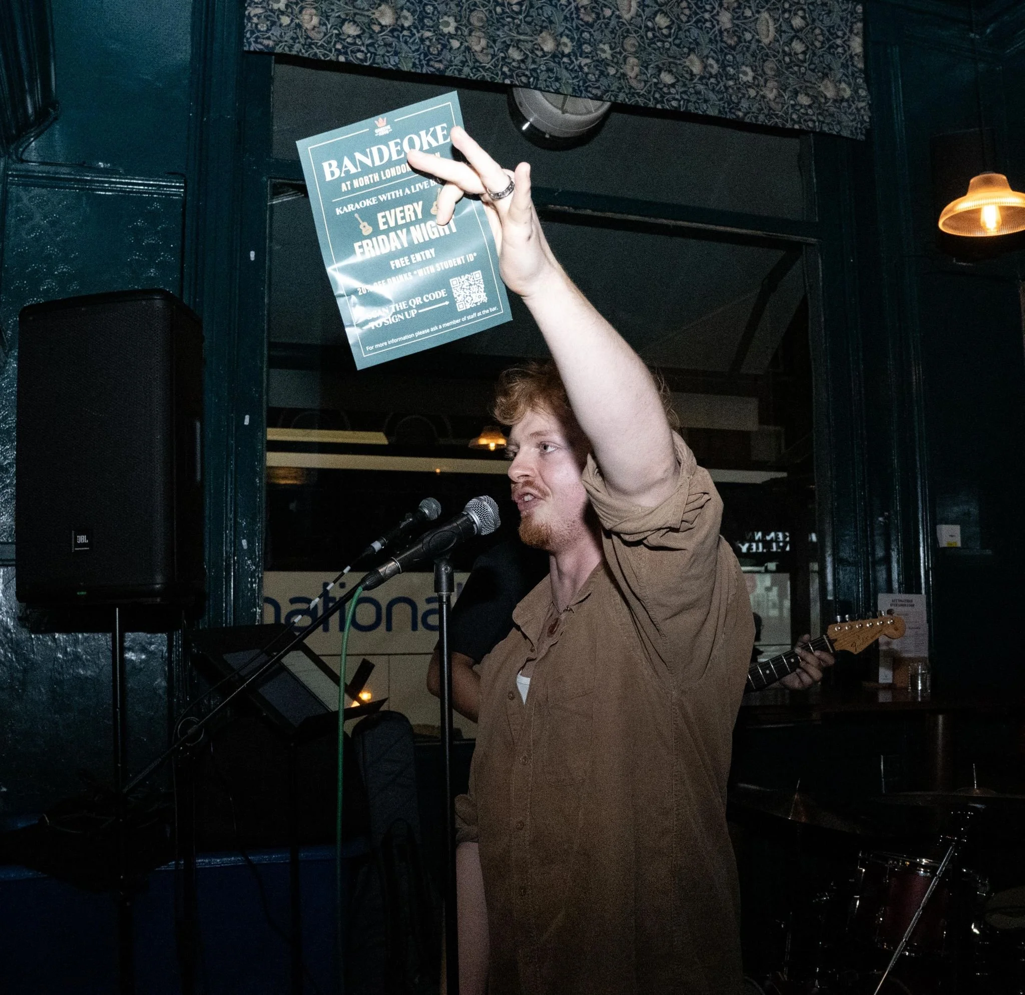 Person holding a promotional flyer for a karaoke event at North London, with stage equipment and a person playing a guitar in the background.