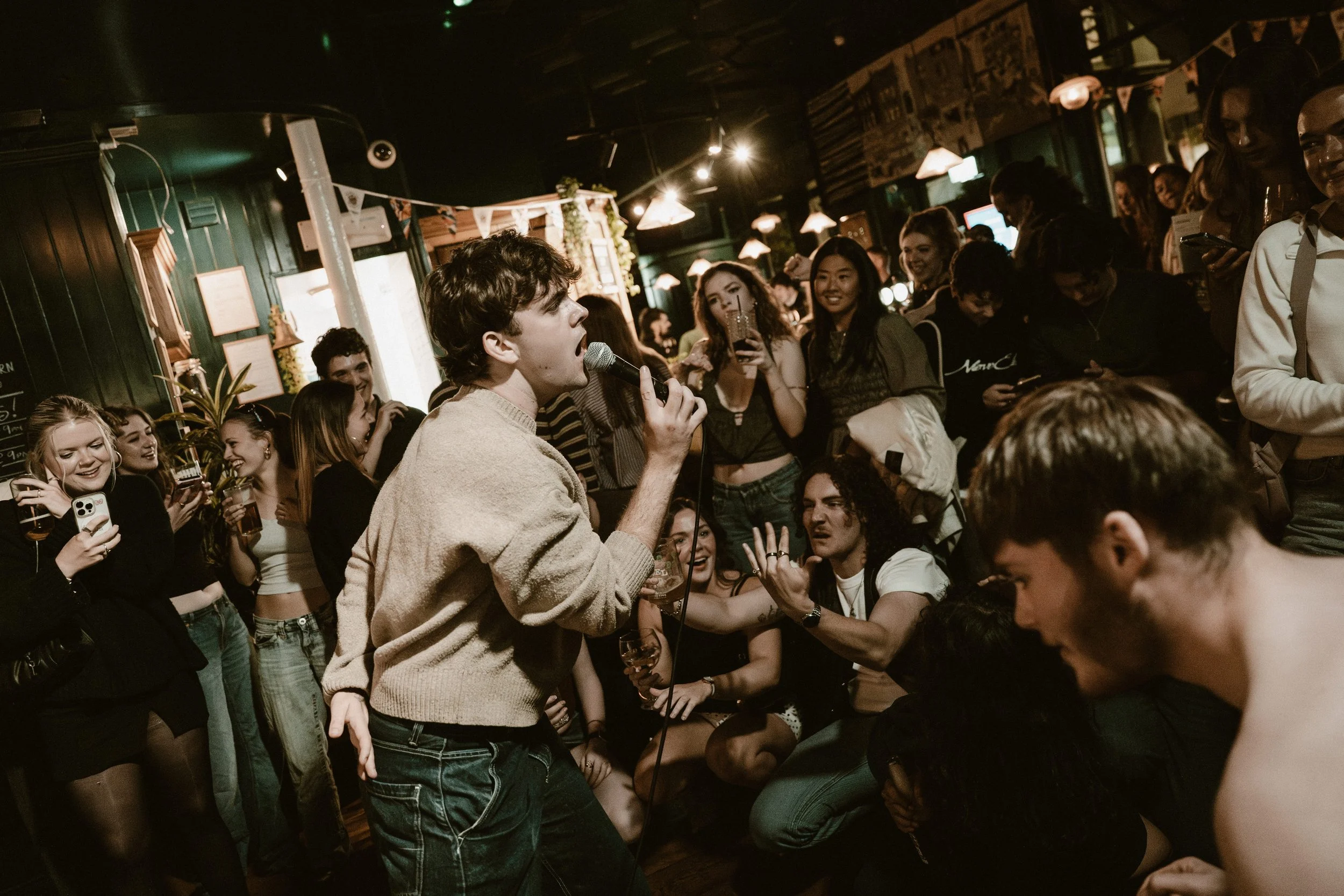 Young man singing into a microphone at a crowded social gathering in a dimly lit bar or pub, with people enjoying drinks and talking.