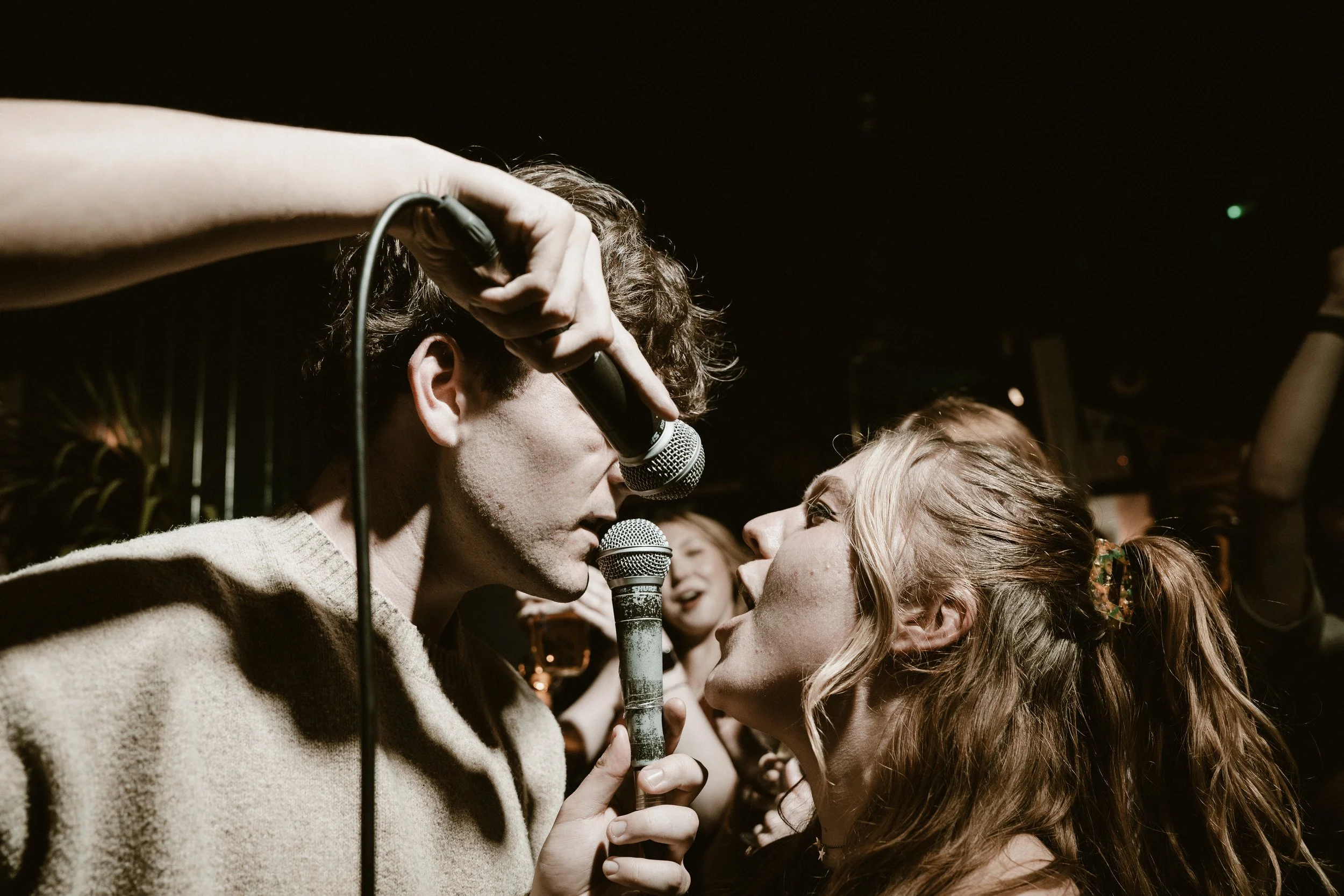 Two young people in an intimate moment, singing into microphones in a dimly lit setting surrounded by others, possibly at a karaoke or live music event.