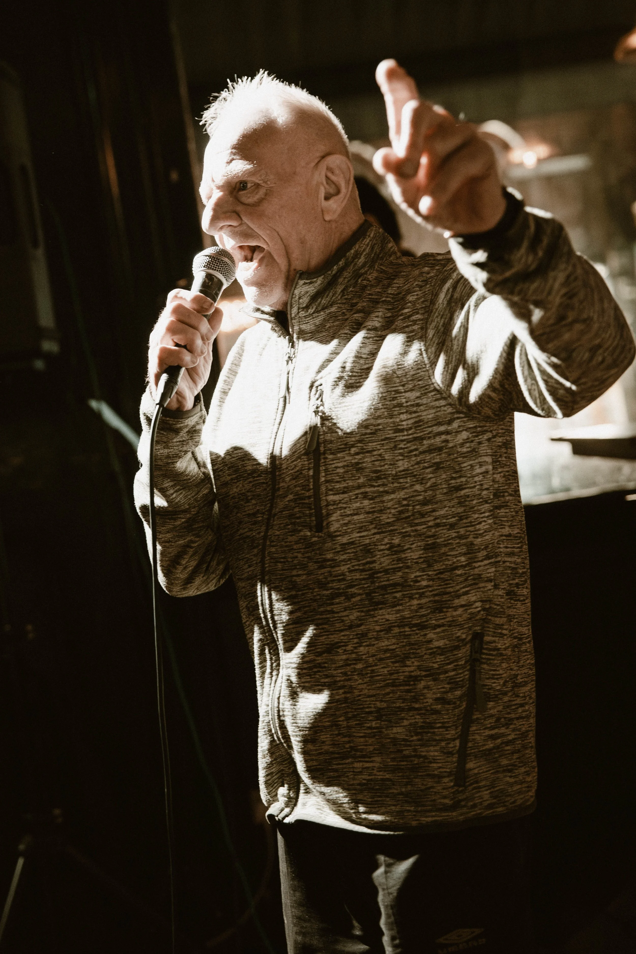 An elderly man with white hair and wrinkles singing into a microphone, pointing upwards, wearing a gray patterned zip-up jacket, in a dimly lit environment.