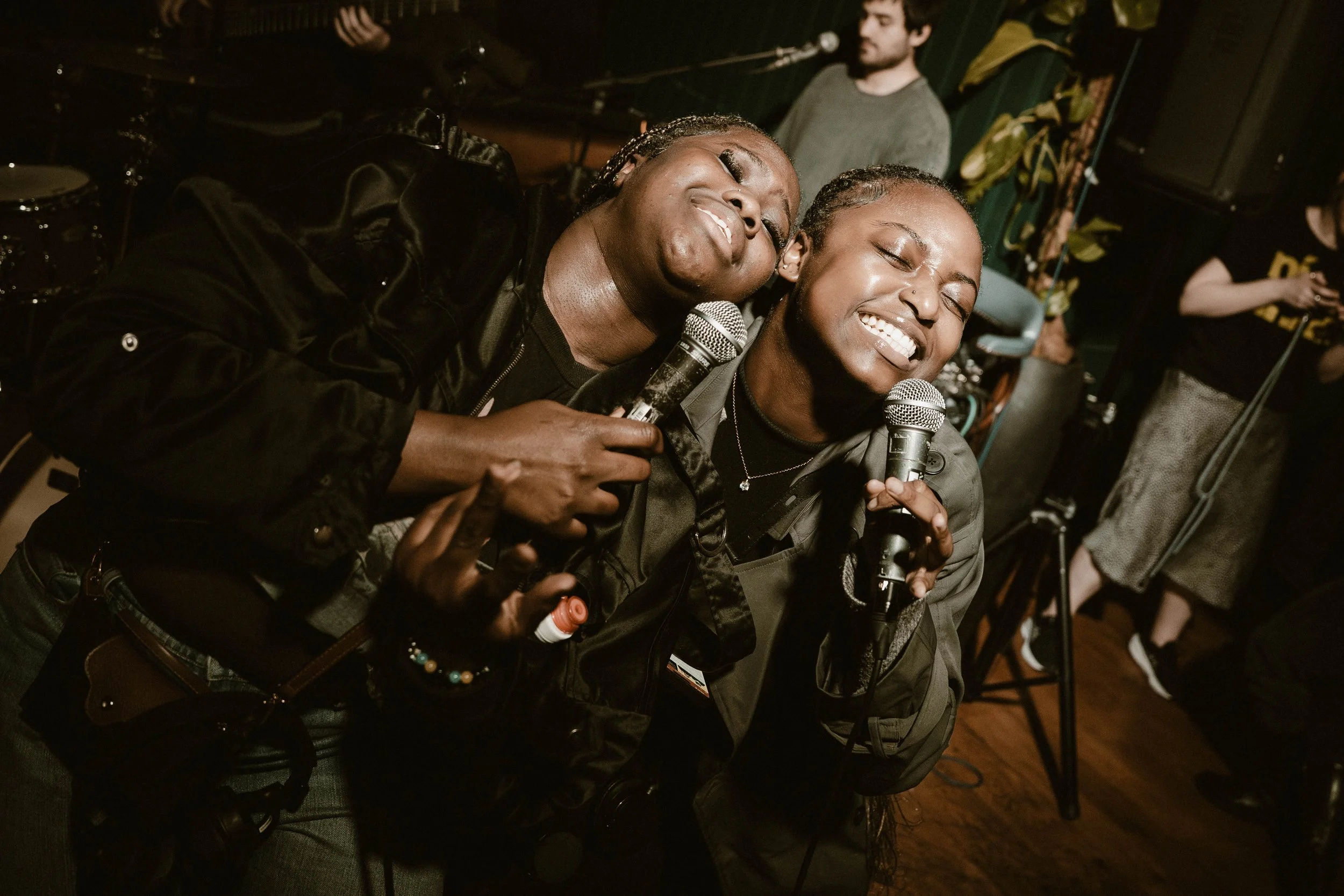 Two women with dark skin smiling and singing into microphones, close to each other, during a live performance or karaoke session in a dimly lit venue.