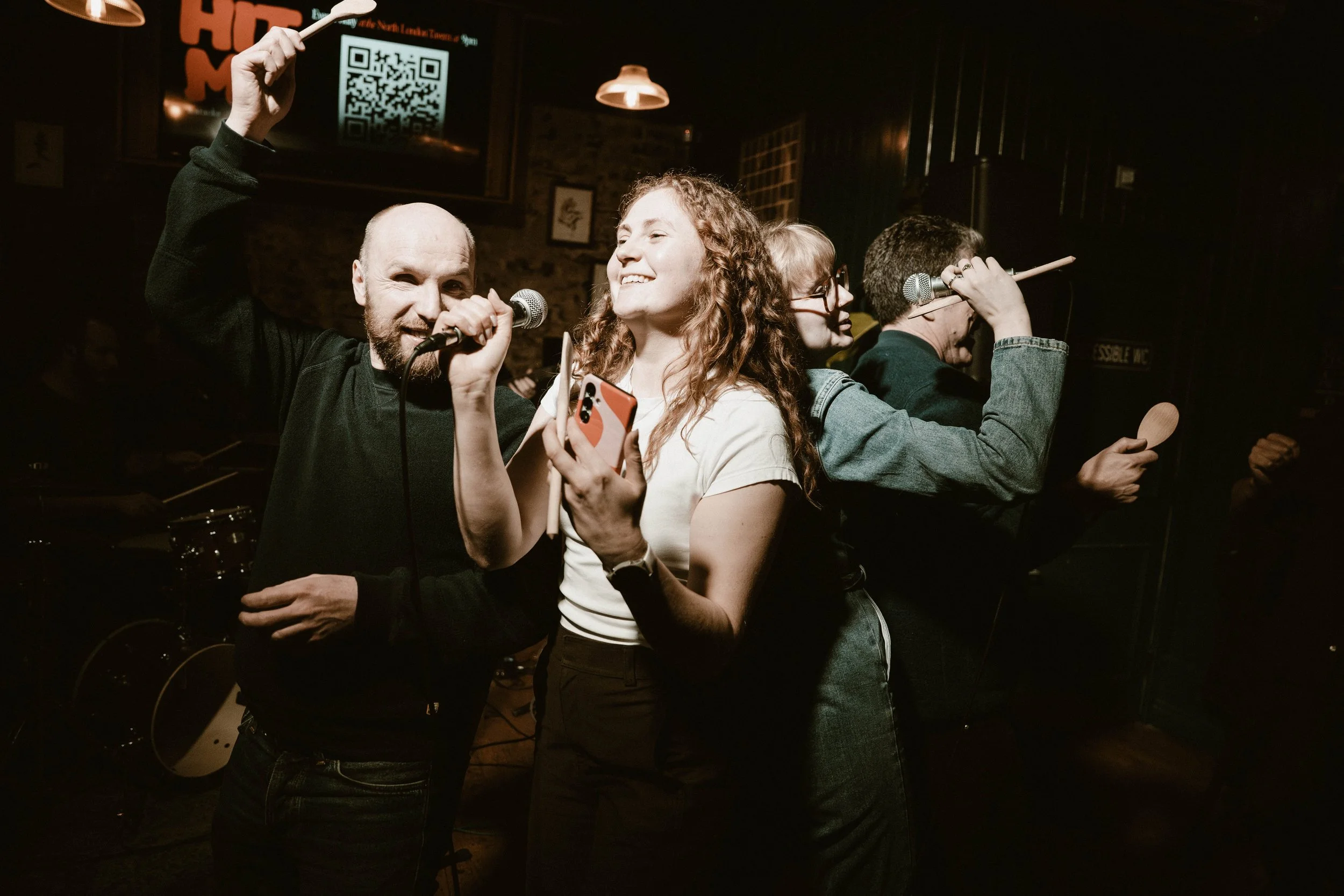 A group of four people performing karaoke in a dimly lit bar. One man with a beard, holding a microphone, is smiling and has his hand raised. A woman with curly red hair is holding a phone and smiling. Two others, one with glasses and short hair, and another with their back turned, are also holding microphones and seem to be singing.