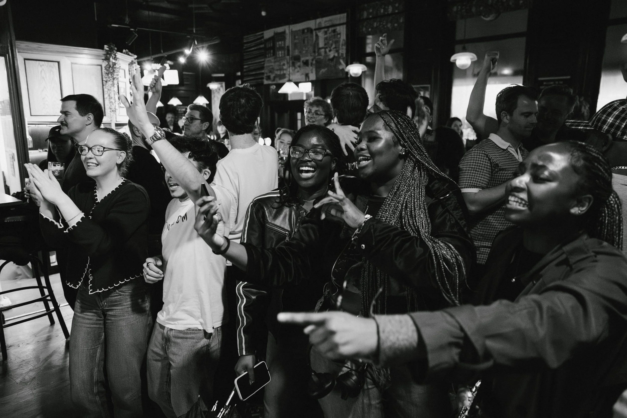 A group of diverse people enjoying a social gathering in a dimly lit indoor space, many smiling, laughing, and cheering, some raising their hands or holding drinks.