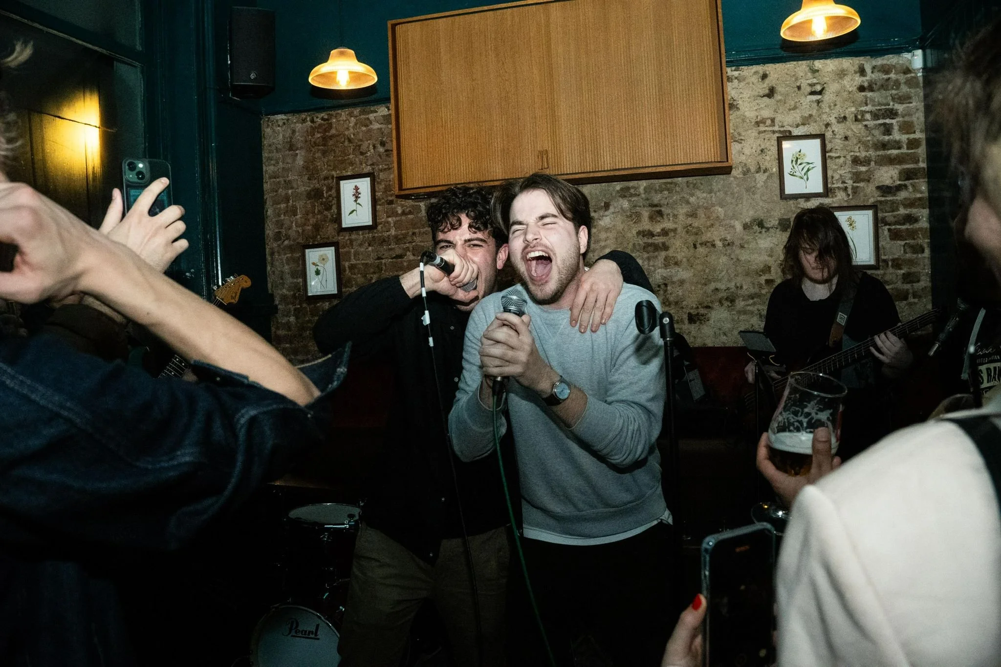 Two young men singing into microphones at a karaoke night, hugging and smiling, with one playing guitar in the background, surrounded by an audience in a cozy brick-walled venue.