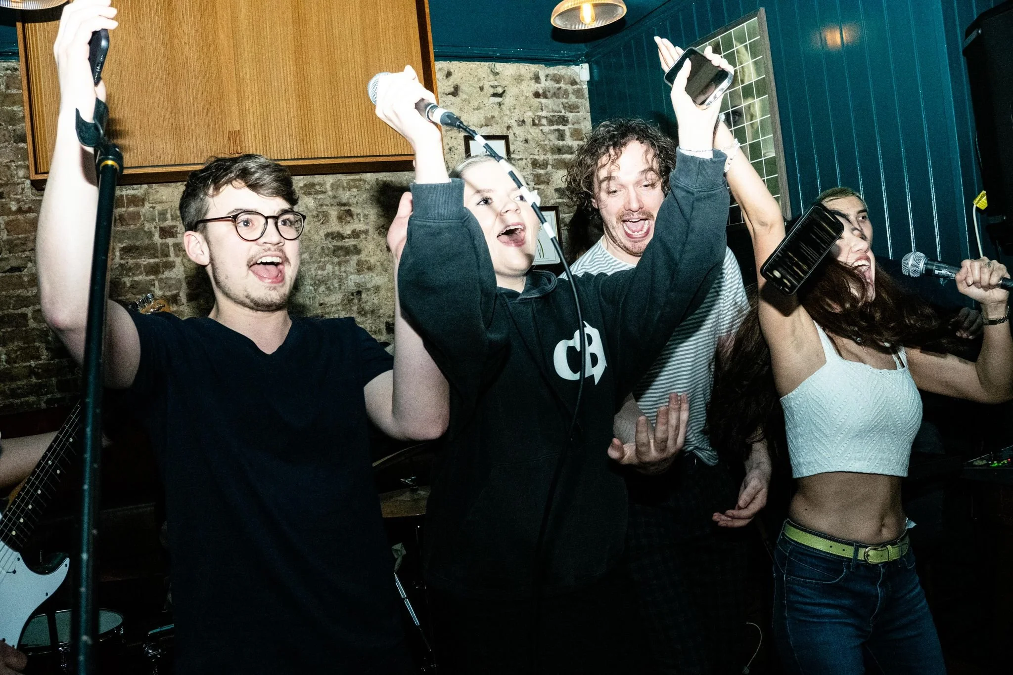 Four young adults singing and raising their hands, with two holding microphones and smartphones, in a dimly lit room with brick walls and ceiling lights.