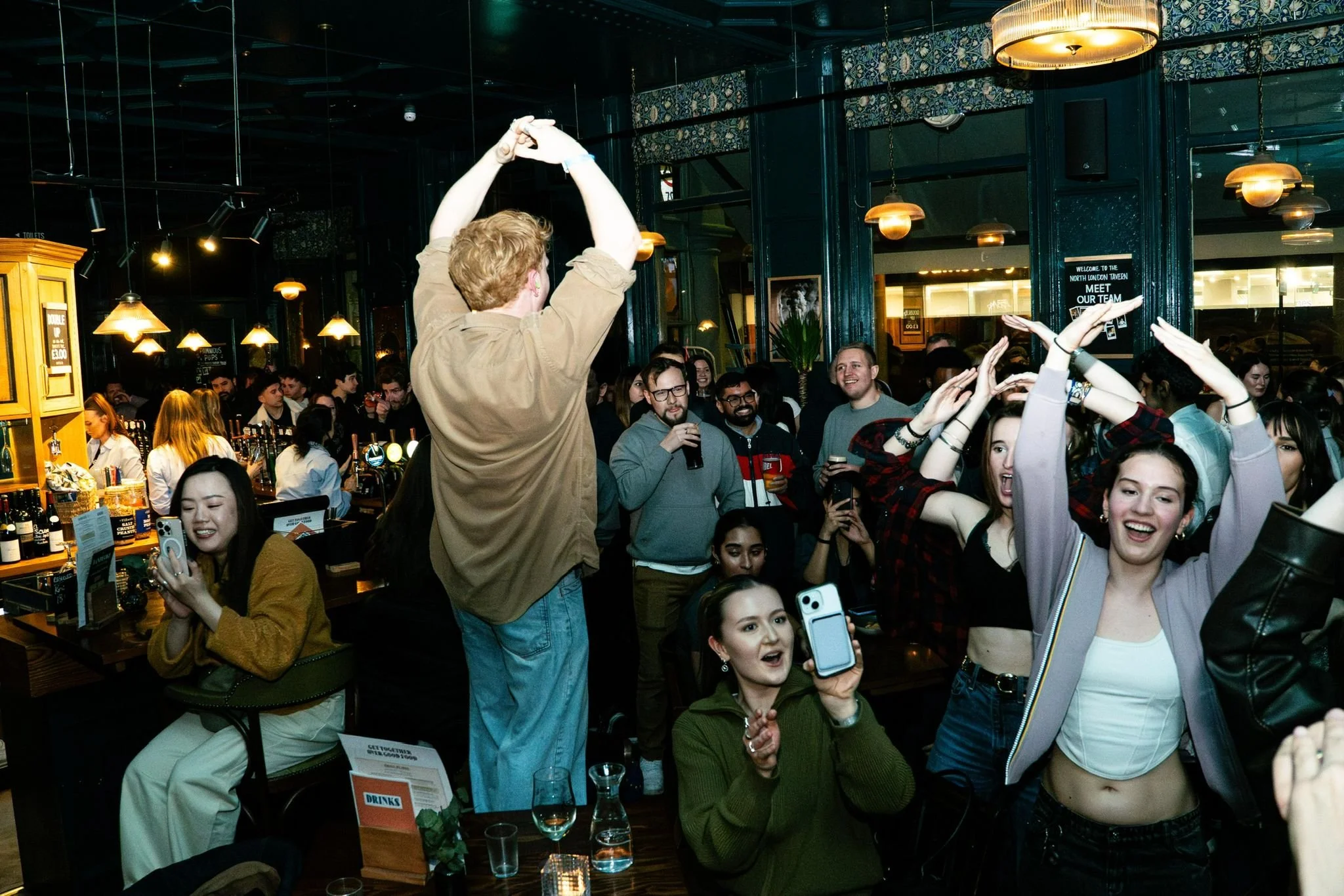 Group of people at a lively social event or party inside a bar or restaurant, with some individuals holding drinks and others using their phones, while a man in a tan shirt is dancing with his arms raised.