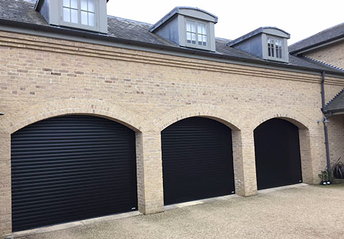 Three closed black garage doors set in a brick building with arched openings and dormer windows.